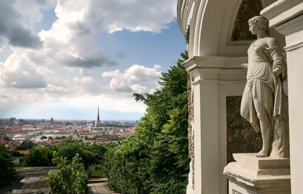 In the image, a close-up of a marble statue inserted into the exterior facade of a building. In the background, a panoramic view of Turin from which the Mole Antonelliana soars